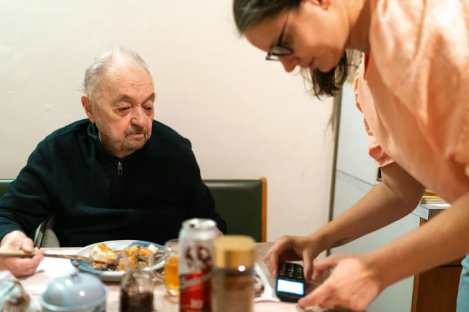 Carer assisting elderly man with digital payment during mealtime at home